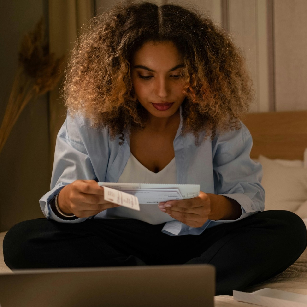 A young woman looks at paper documents while sitting cross-legged on a bed in front of an open laptop