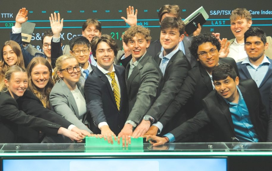 A group of students dressed in business wear cheering. They stand in front of a screen with stock market information.