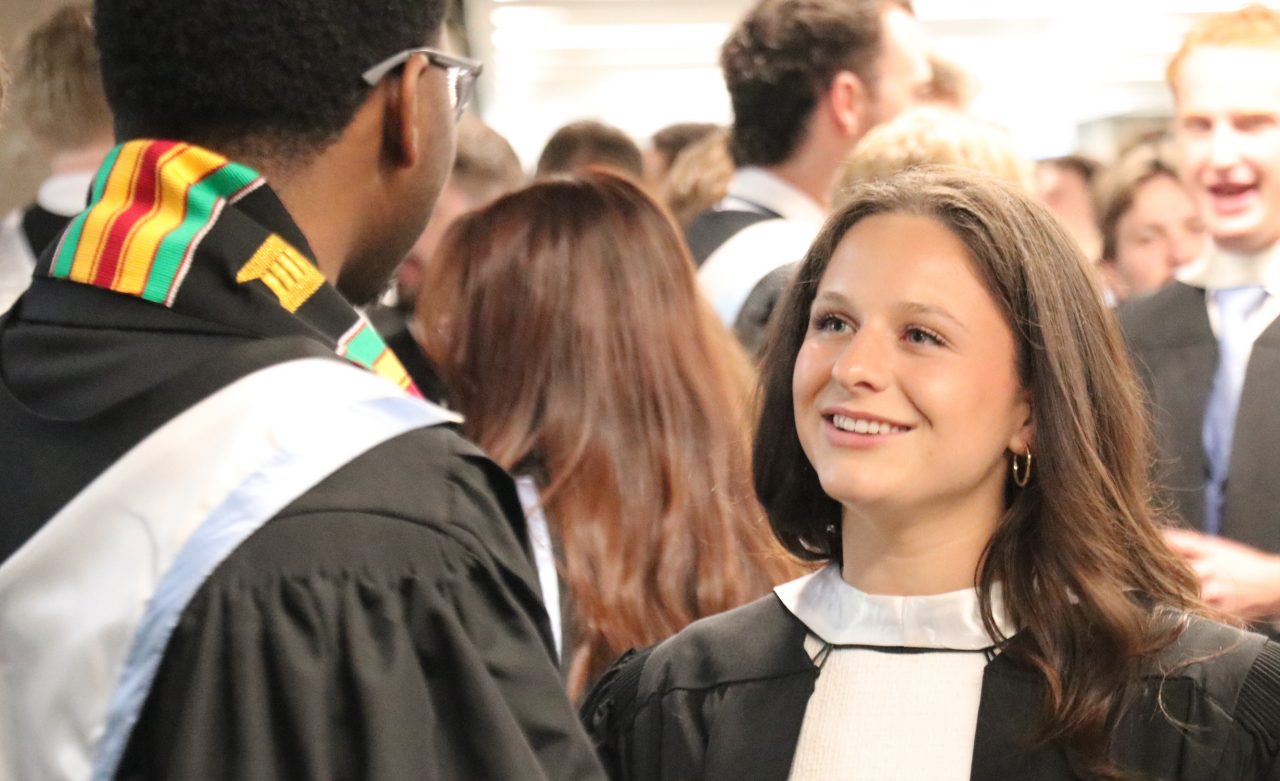 A student smiles at another student (facing away from camera). They are dressed in convcoation wear, with one wearing a kente sash in red, yellow, green and black.