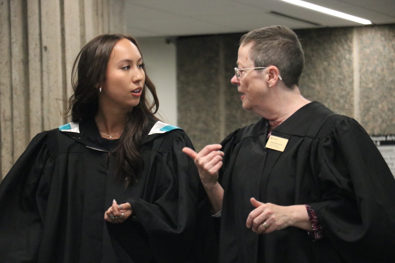 A student and staff member chat animatedly. They are dressed in convocation robes. 