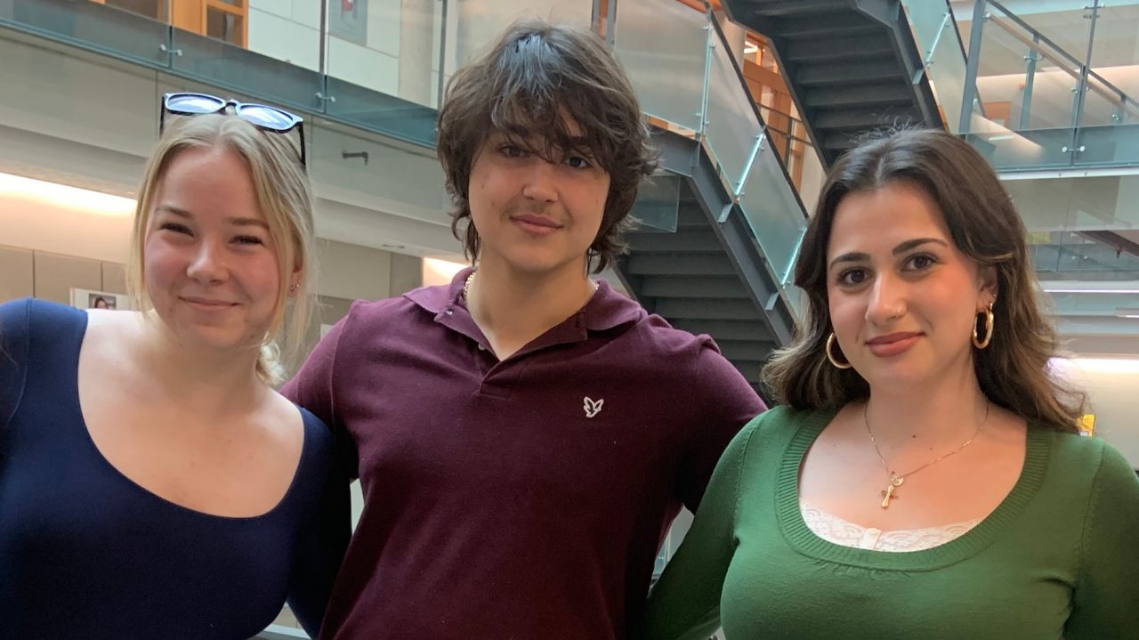 5 students pose, arms around each other and smiling, with a staircase inb the background.