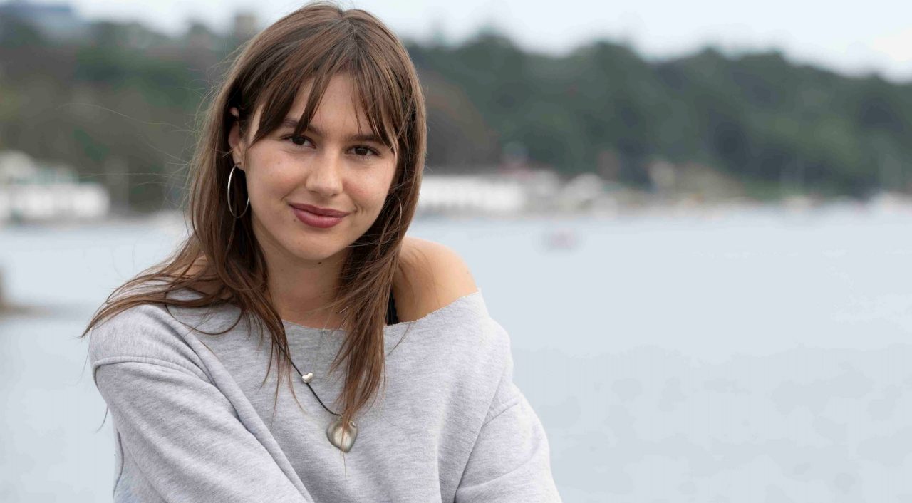 Abby sits on a rock facing the camera, smiling. In the background is water and the coastline. 