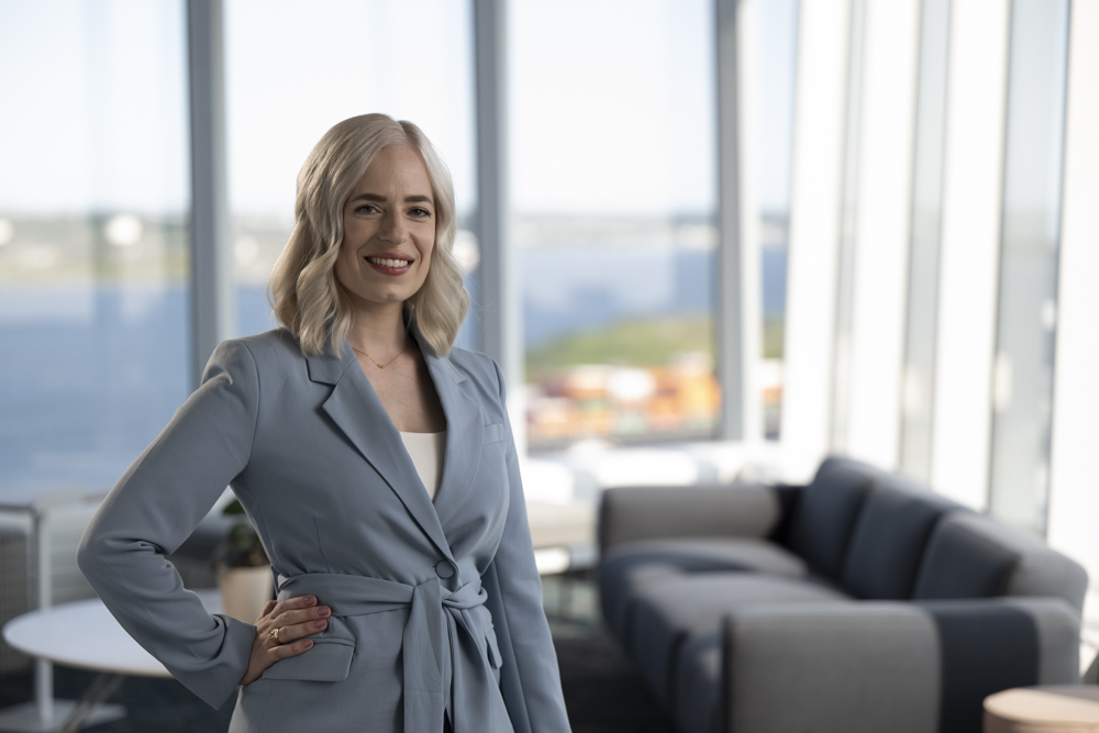 Kristie poses in a blue suit, with a picture window overlooking a harbour behind her.  