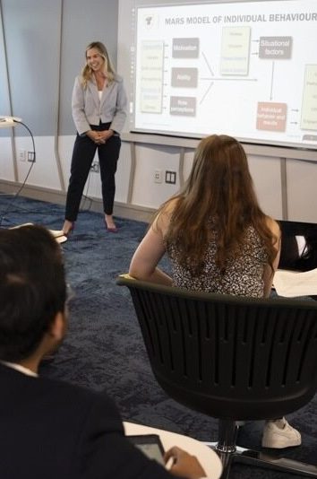 Two students presenting at the front of the room, to a group of classmates sitting in chairs. 