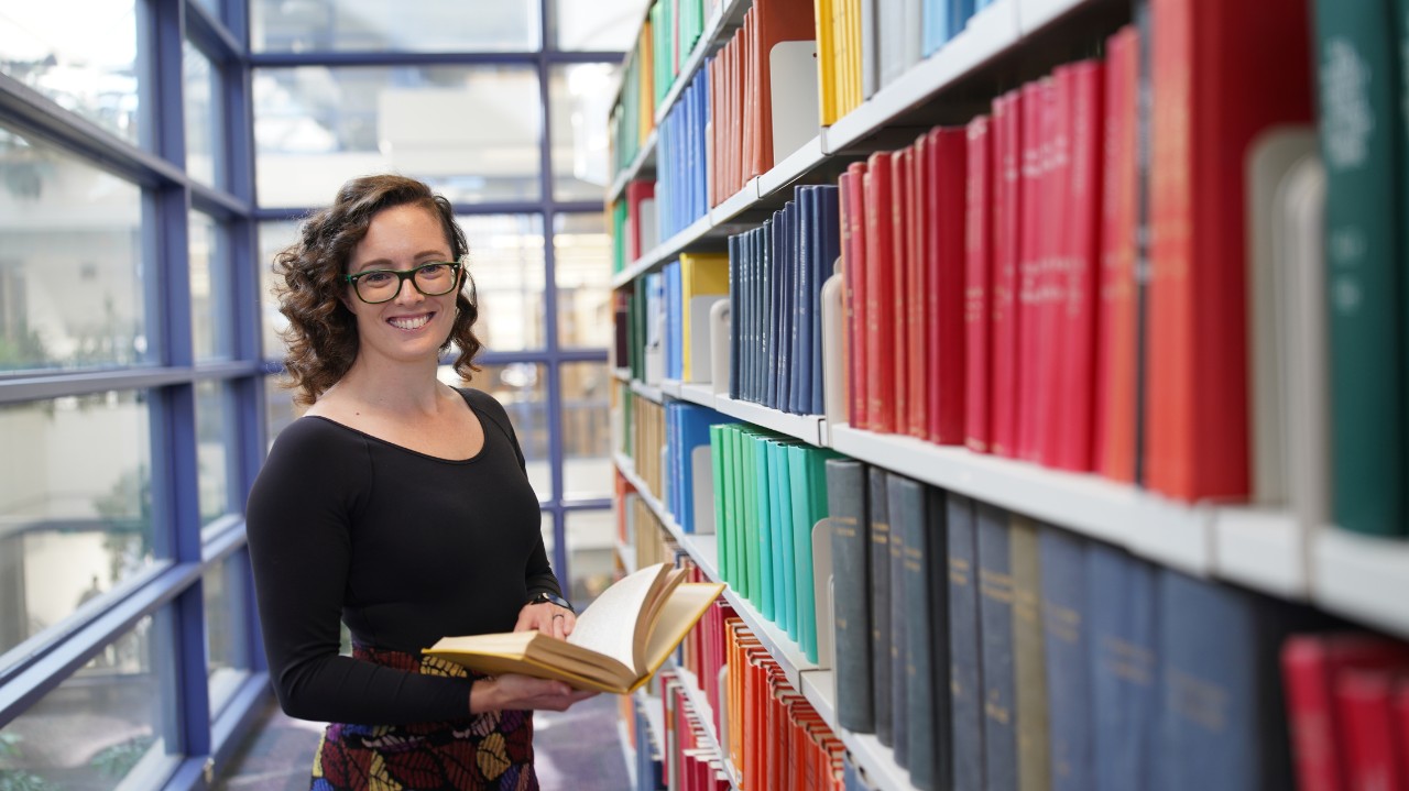 A person wearing a black shirt stands next to shelves filled with books, holding a book and smiling.