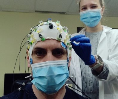 A professor wearing a cap with electrodes connected to it. A student wearing gloves behind him. 