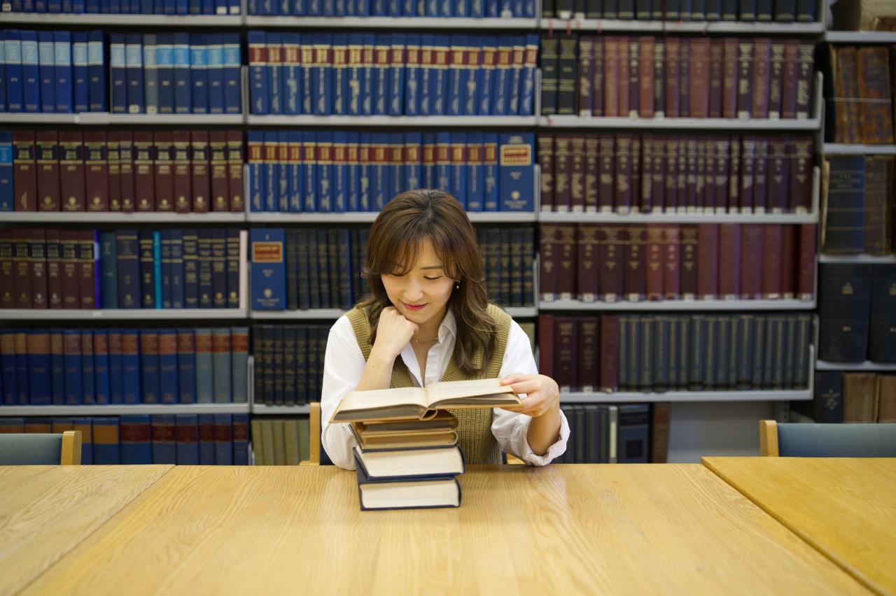 A person wearing a white shirt and brown sweater sits at a table reading a book in front of shelves filled with books.