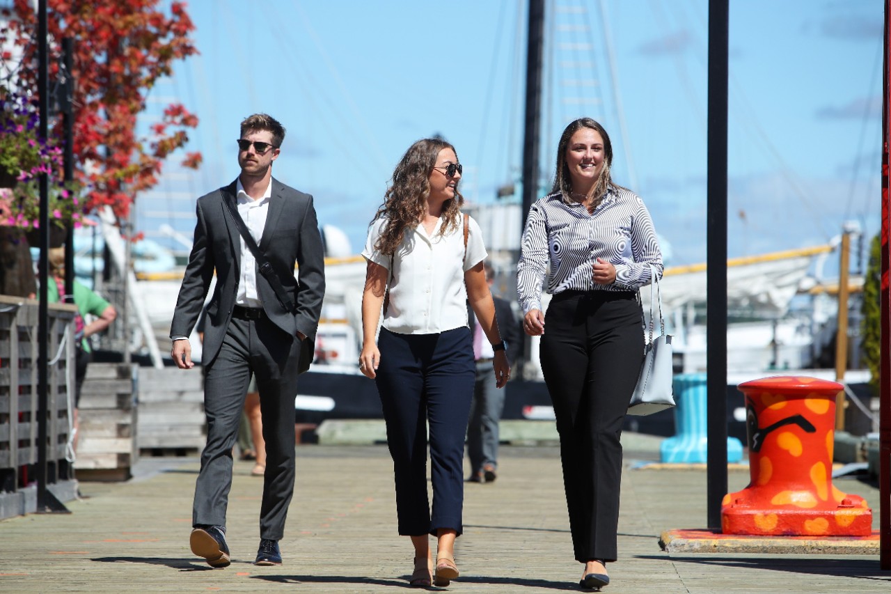 Three CRMBA students wearing professional attire walk along the Halifax waterfront on a sunny day.