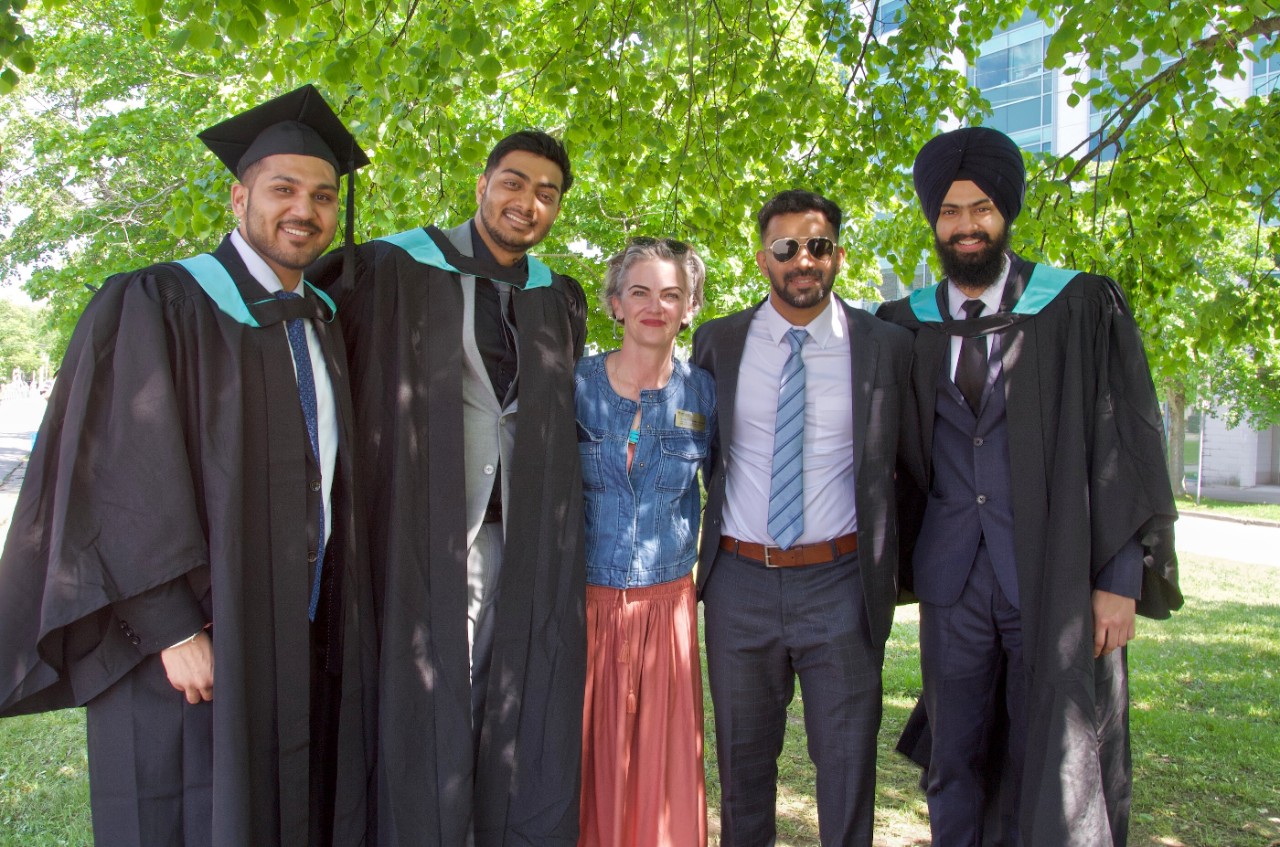 Four students in convocation dress stand with a staff advisor under a tree, smiling.  