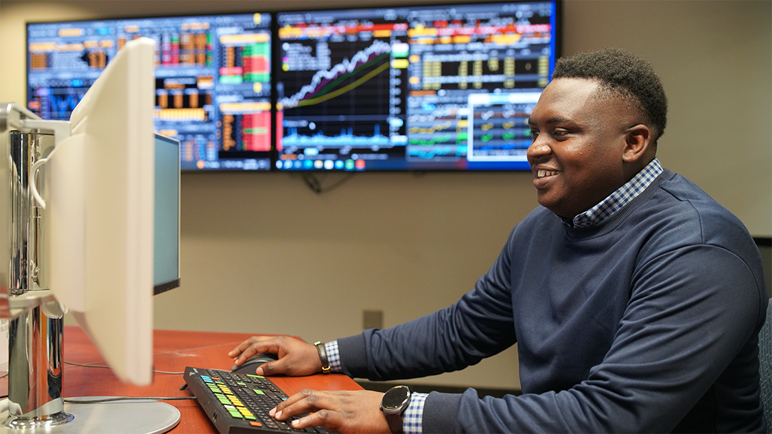 A student sits at a computer with a multi-coloured keyboard. Screens in the background show tables and charts.  