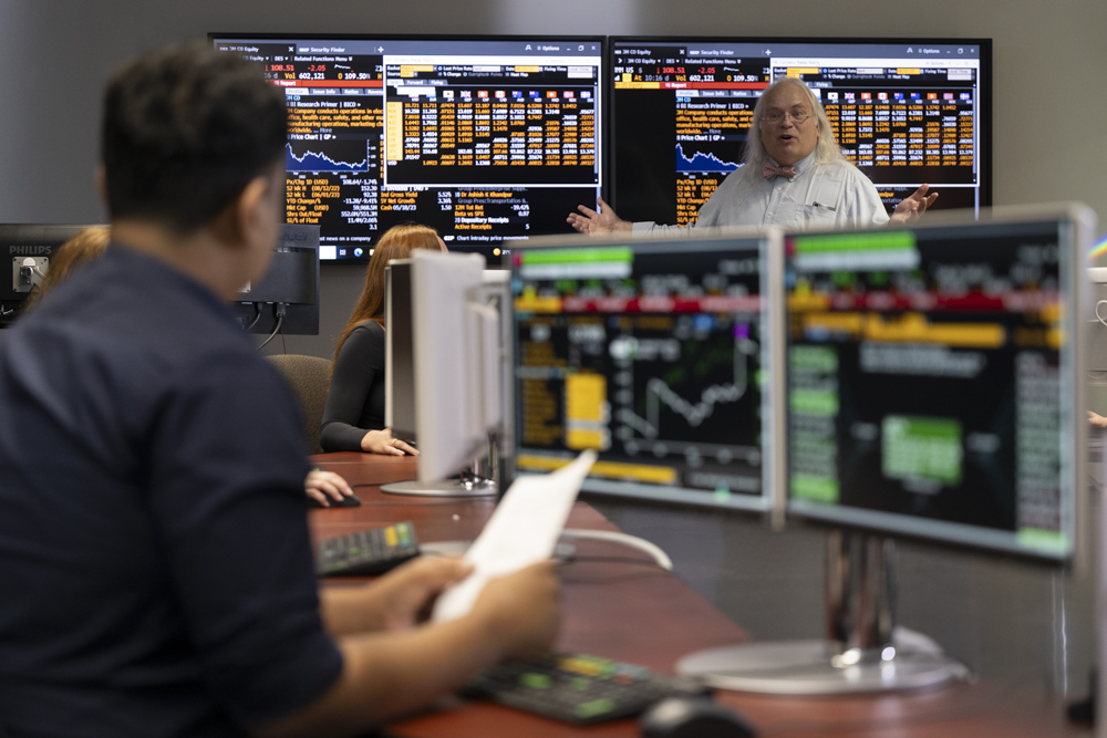 A student looks up from dual monitors showing colourful graphs to a professor explaining stock information on a large screen. 