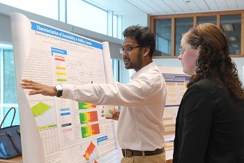 Two people stand in front of a large white poster containing text and visuals from a research project. 