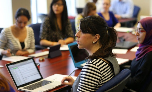 Six people sit around a large boardroom-style table with laptops or tablets in front of them looking left at a speaker off-screen.