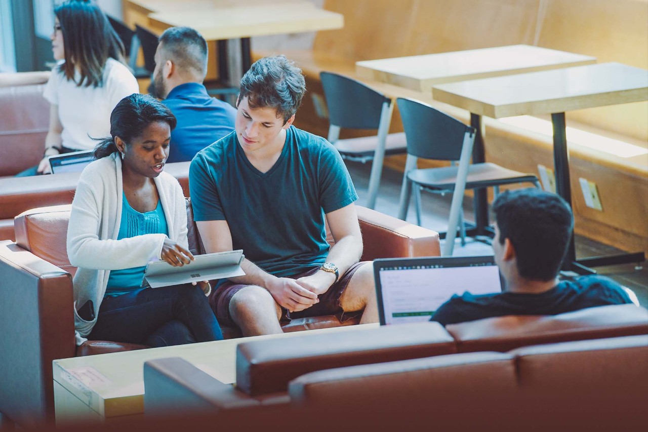 Students sit in a light-filled atrium, reviewing something on a tablet. 