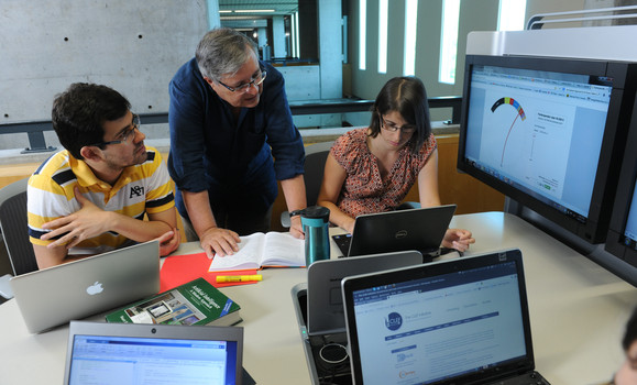 Two people sit at a desk looking at a laptop and another person stands between them looking at the laptop and talking.