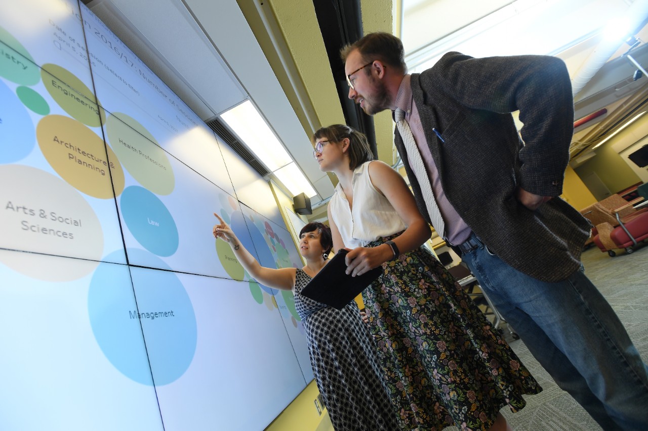 Three students stand in front of a large screen, gesturing to clusters of dots representing data that they are discussing. 