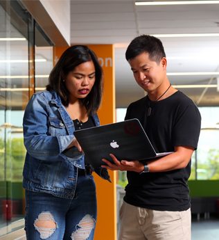two indigenous students standing outside of a room and looking at a laptop together