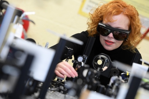 A woman with red hair wears dark safety glasses while operating lab machinery.