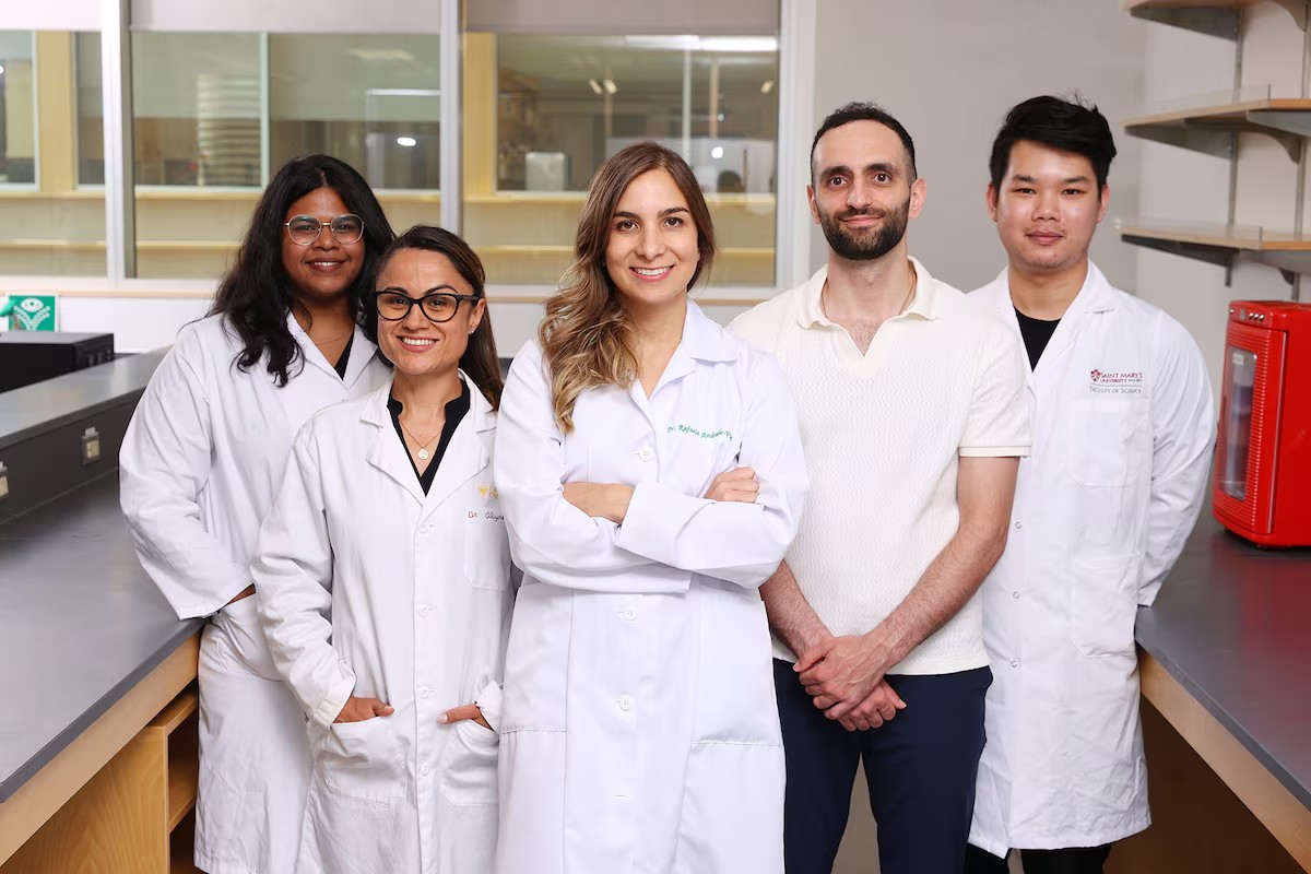 Andrade stands with arms crossed smiling with a team of four others, all are wearing white lab coats.