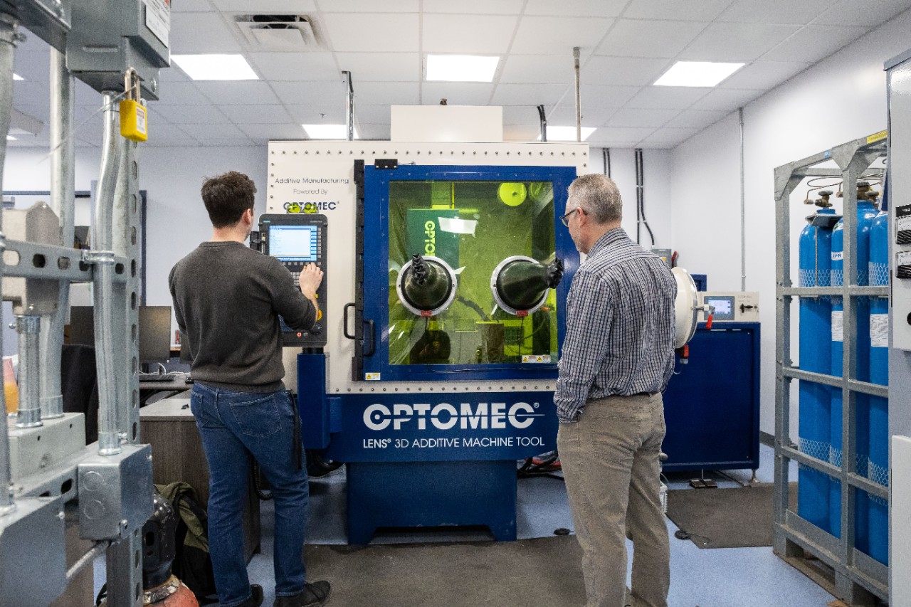 Two individuals standing near a machine labeled "Optomec".