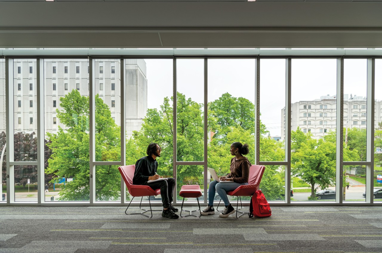 Two students sit by a wall of windows