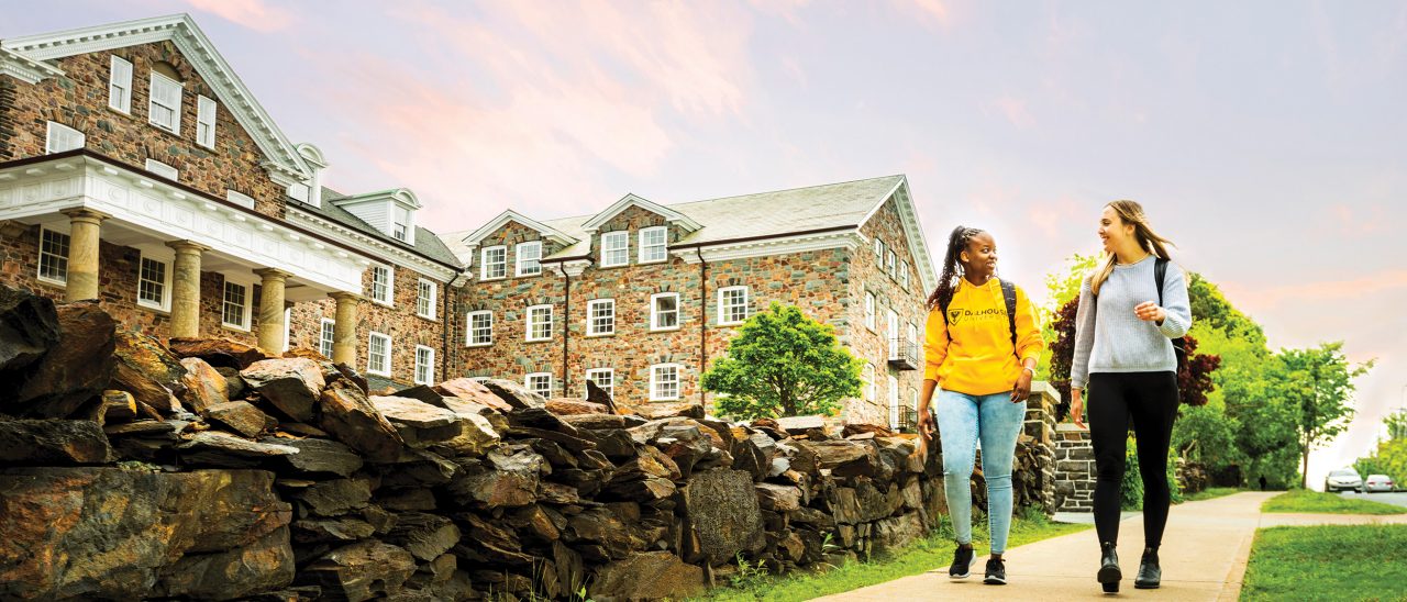 Two female students walk on the sidewalk in front of old stone buildings at sunset. The sky is blue with pink clouds.