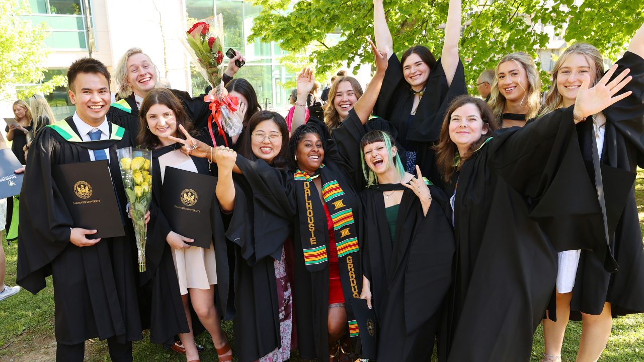 A dozen students are wearing their graduation gowns with their arms up in the air in celebration. 
