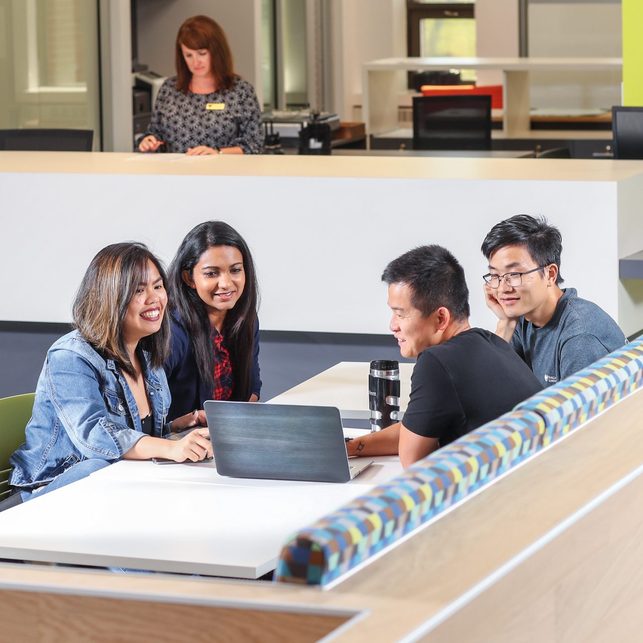 Four students sit at a table looking at a laptop, while a librarian works in the background.