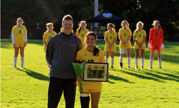 Jill Lockerby standing on a soccer pitch holding a frame and flowers