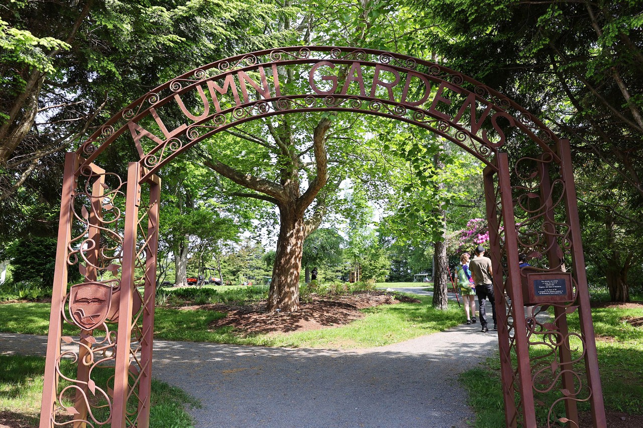 An ornate copper gate with the words "Alumni Gardens" across the top leads to a leafy, green garden full of trees.