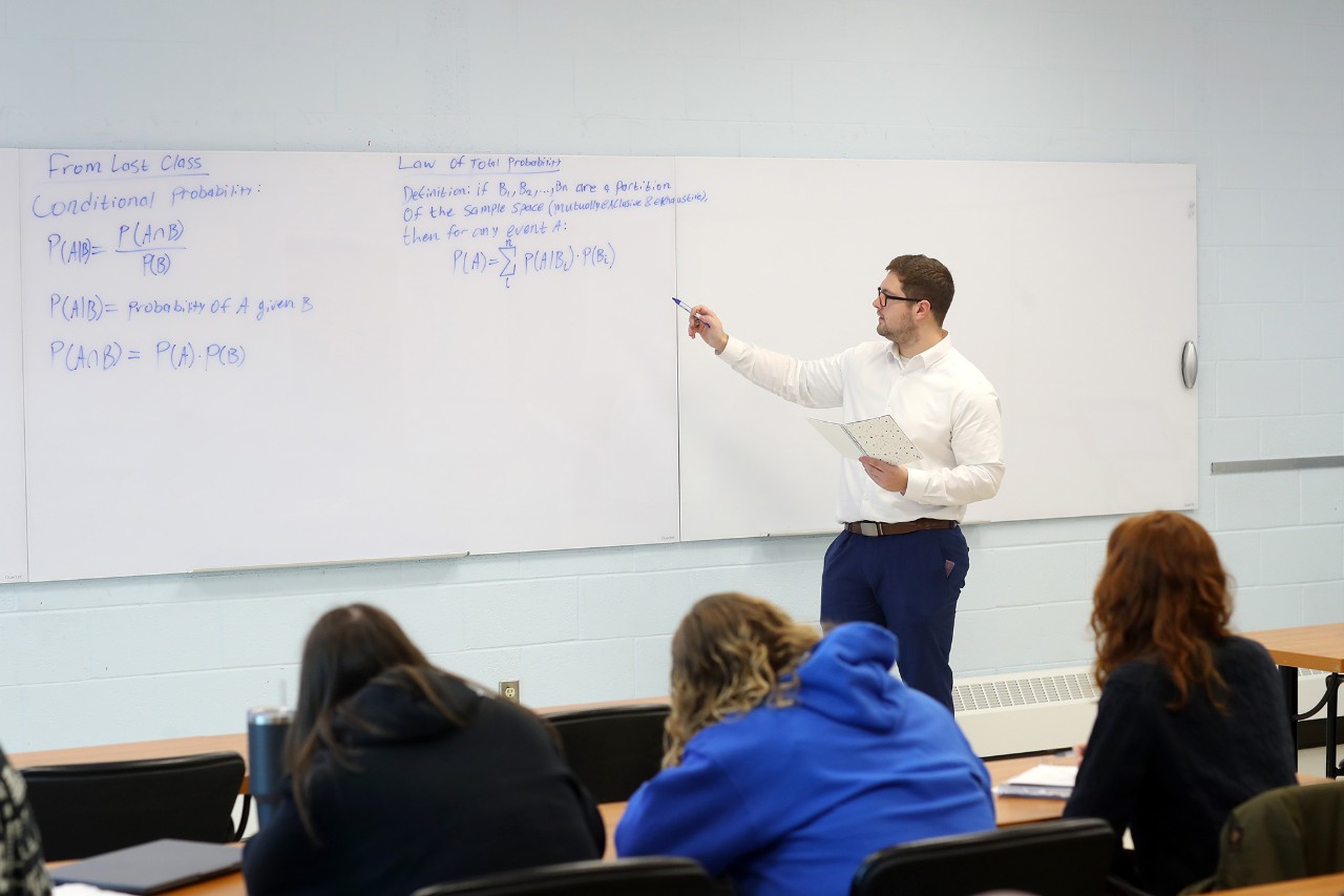 Male class instructor pointing at blue writing on a whiteboard. Students sit in the foreground.