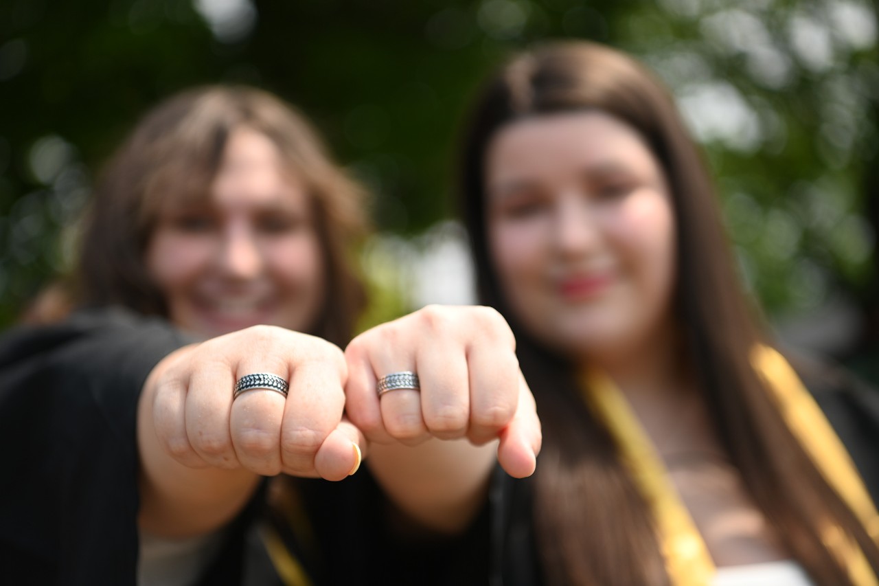 Two students wearing convocation robes raising their barley rings to the camera.