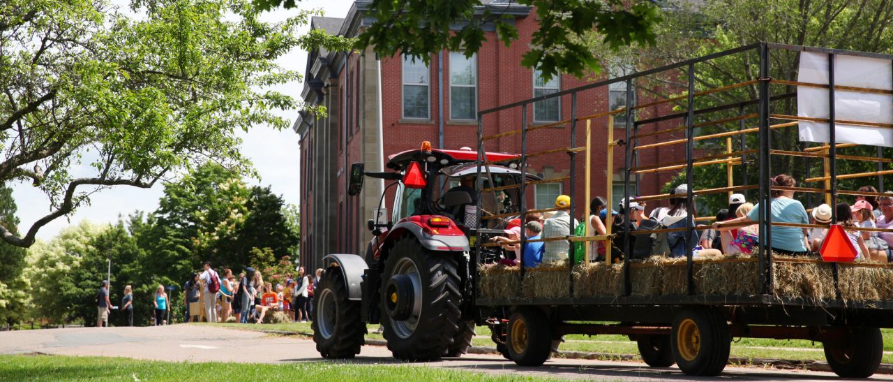 A red tractor pulling a flatbed filled with guests sitting on hay bales approaches a brick building.