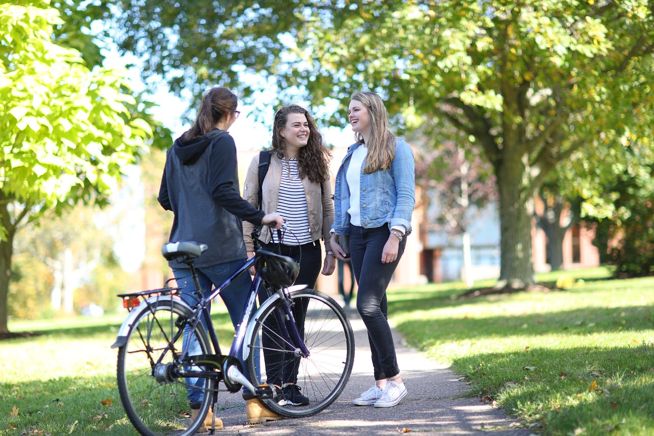 Three students standing on a path talking with each other. Two students face the camera. The student with their back to the camera is holding a black retro bike.