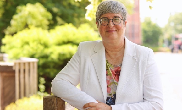 Dr. Heather Bruce smiles at the camera. She is wearing a white blazer and glasses. Greenery of the Agricultural Campus is blurred in the background.