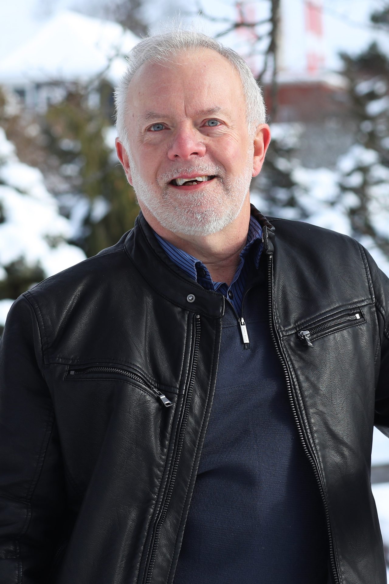 Smiling man with short white hair wearing black leather jacket, pictured on a snow covered Agricultural Campus, with trees and the iconic watertower in the background.