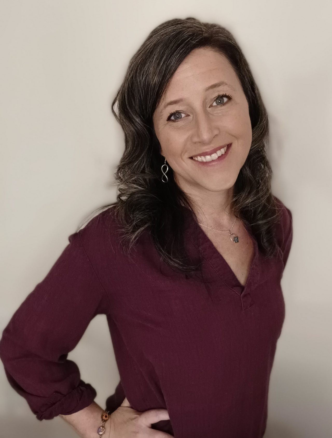 Smiling woman with long dark hair wearing burgundy top poses with her hand on her hip.