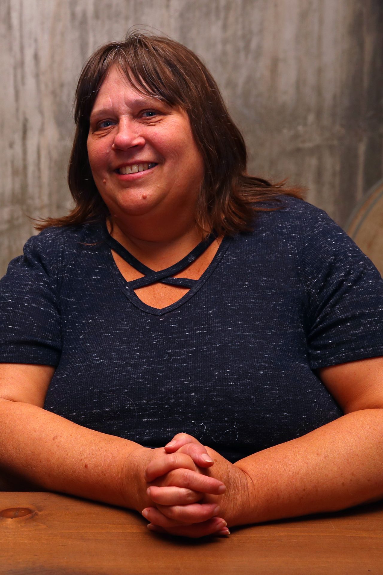 Smiling woman with long dark hair wearing a blue shirt sitting a table with her hands clasped in front of her.