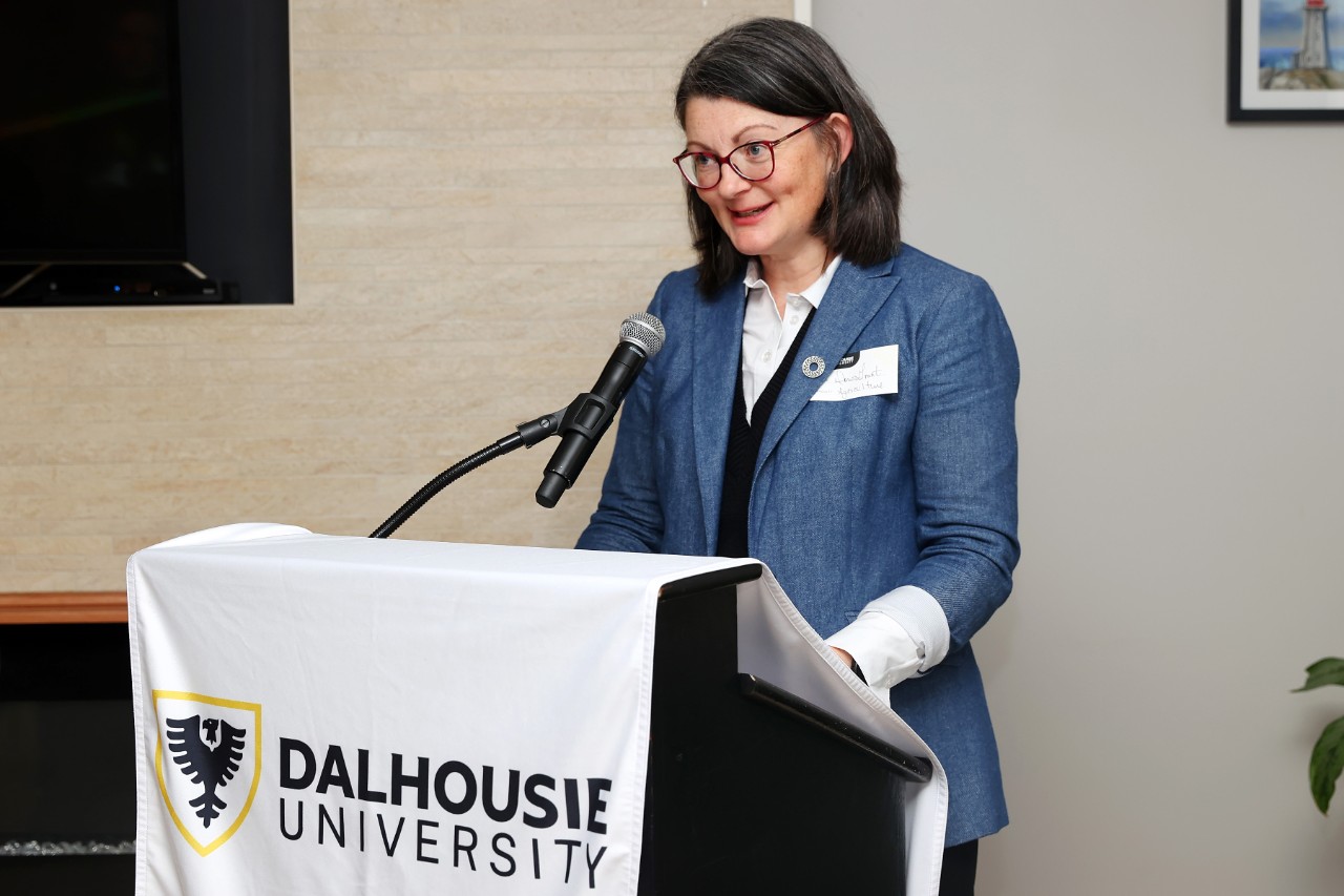 A woman with dark hair wearing glasses stands at a podium with a microphone. The podium is draped with a "Dalhousie University" banner.