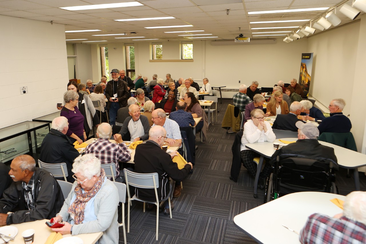 Groups of older alumni sit chatting at tables in large room.