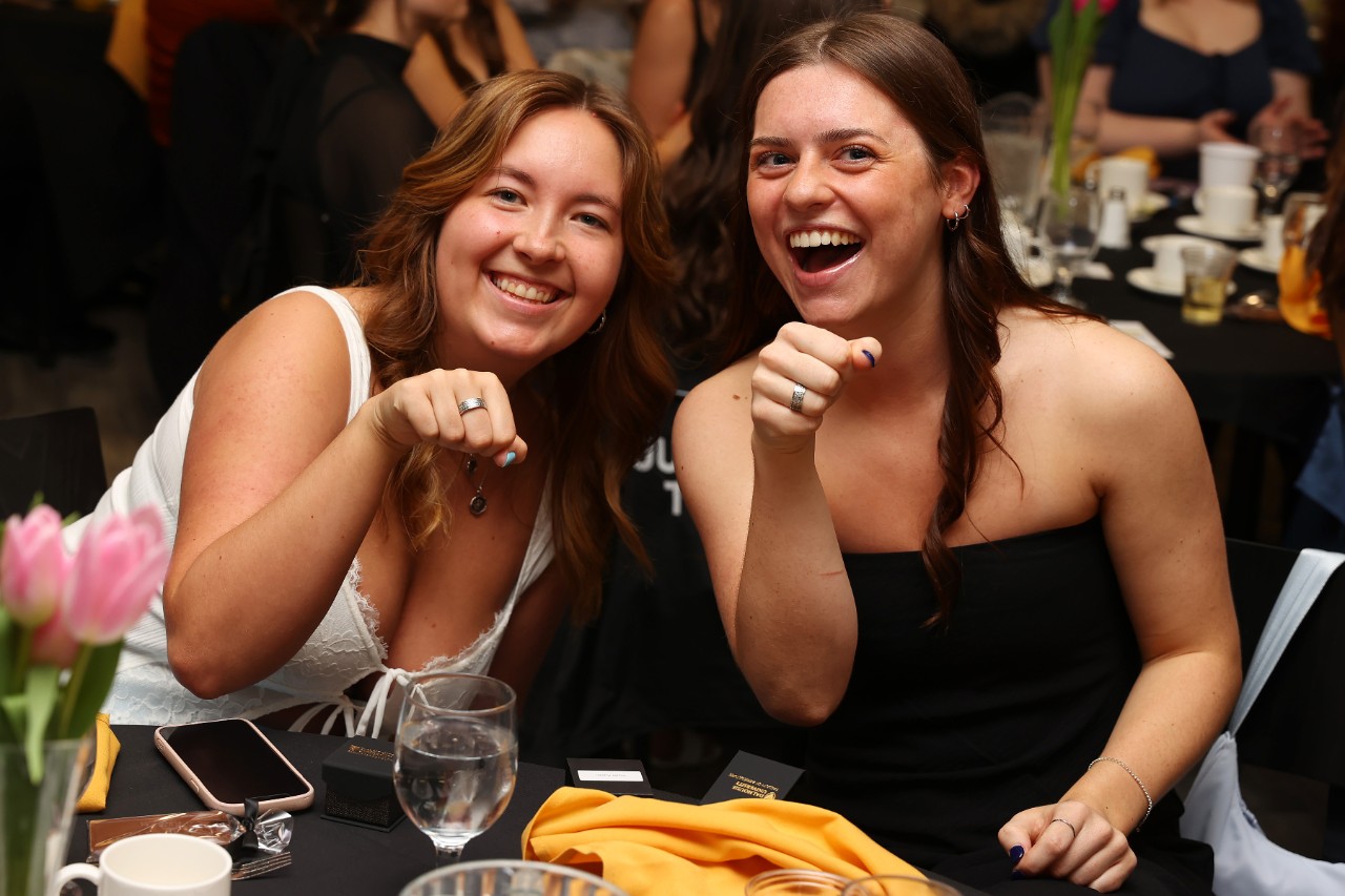 Two students sit at a table smiling, with their fists raised to display their Barley Rings.