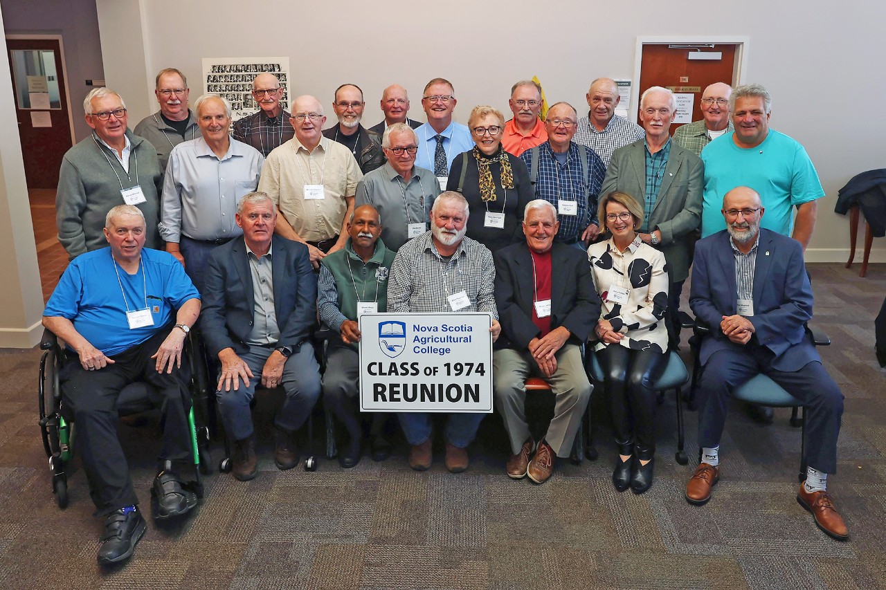 A group of older individuals stand in three rows. The individuals in the front row are sitting in chairs, with one holding a sign reading "Class of 1974 reunion."