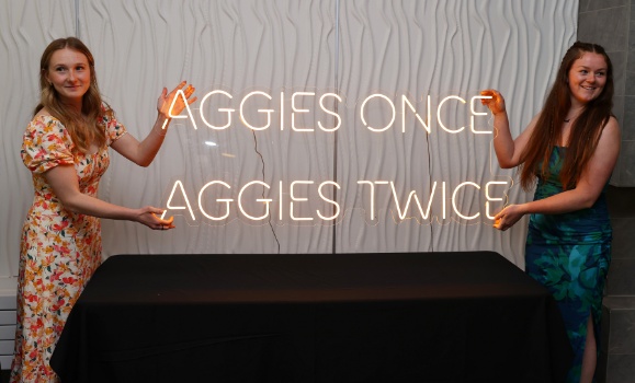 Two Agriculture grads holding an "Aggies once, Aggies twice" neon sign.