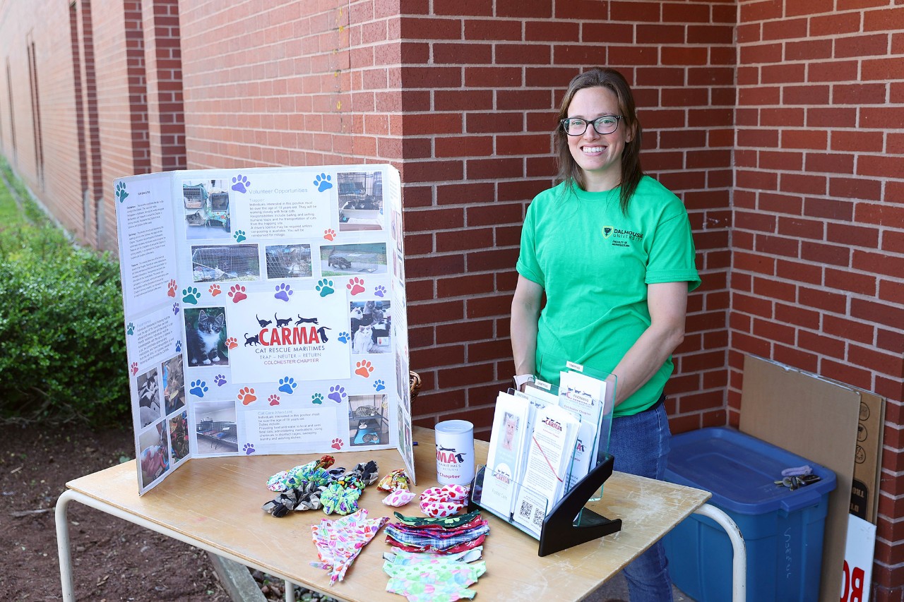 Woman with long dark hair and glasses wearing a bright green shirt stands behind a table displaying posters and brochures.