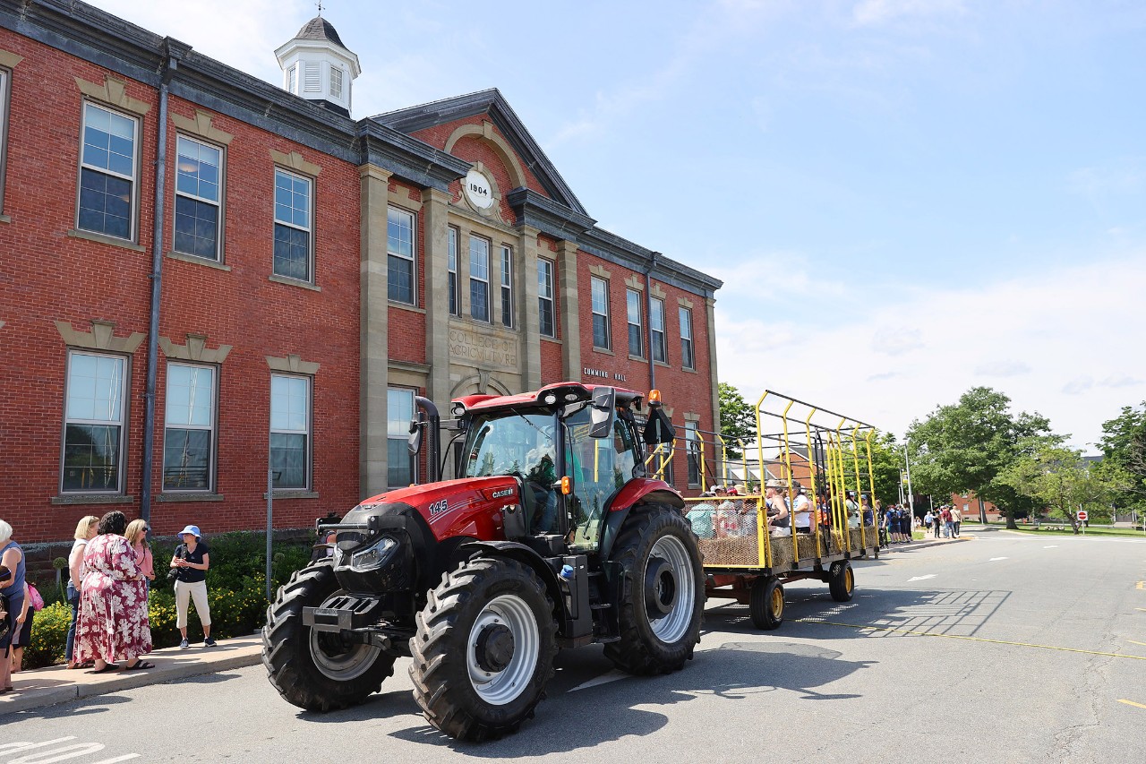 A red case tractor pulling a wagon full of riders in front of Cumming Hall - a red brick building with a white tower.