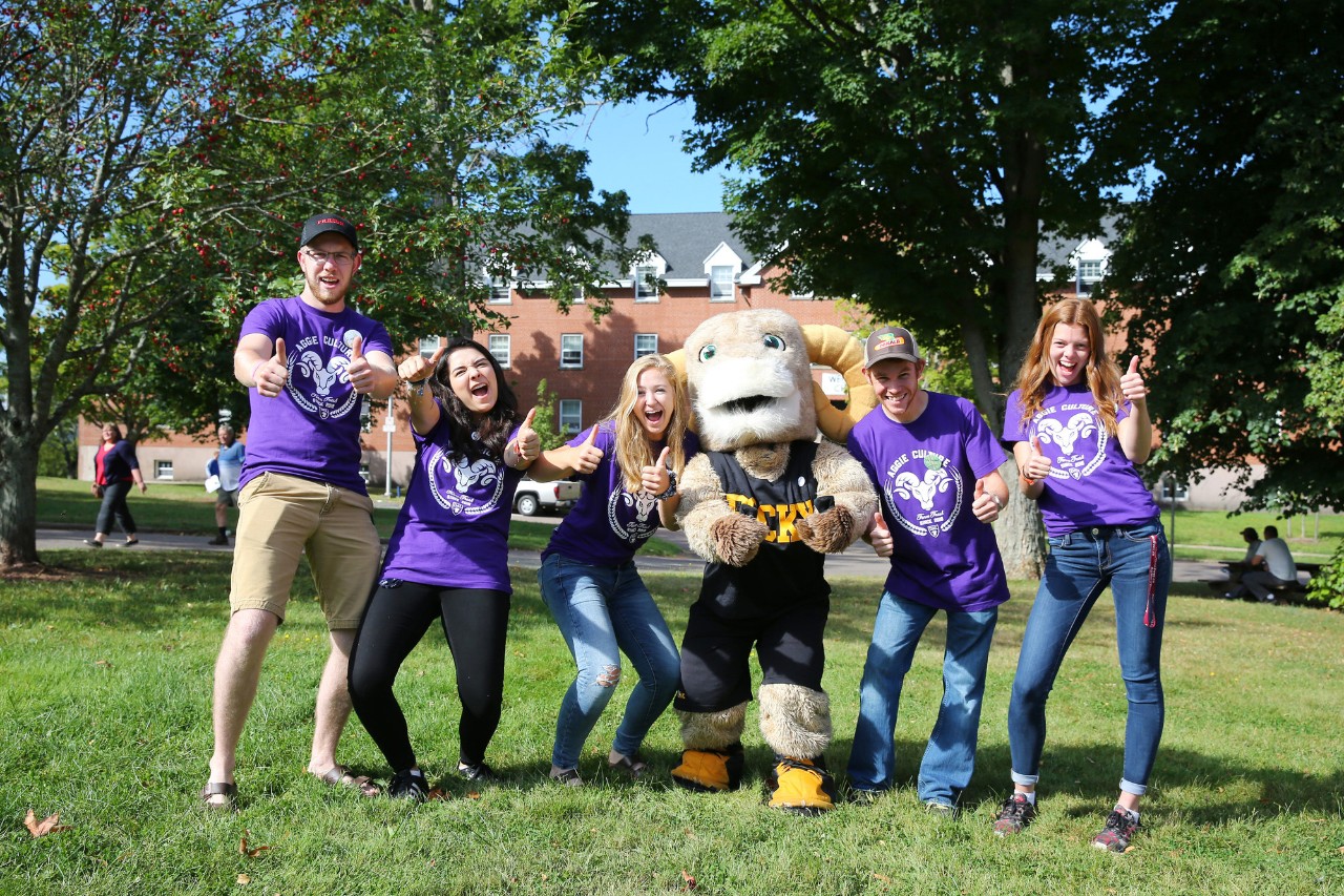 Five students wearing purple t-shirts enthusiastically giving thumbs up, with Rocky mascot standing in the middle.