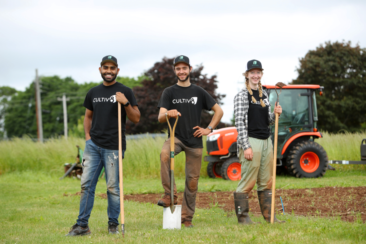 Two men and one woman pose with garden tools in a field with a red tractor visible in the background