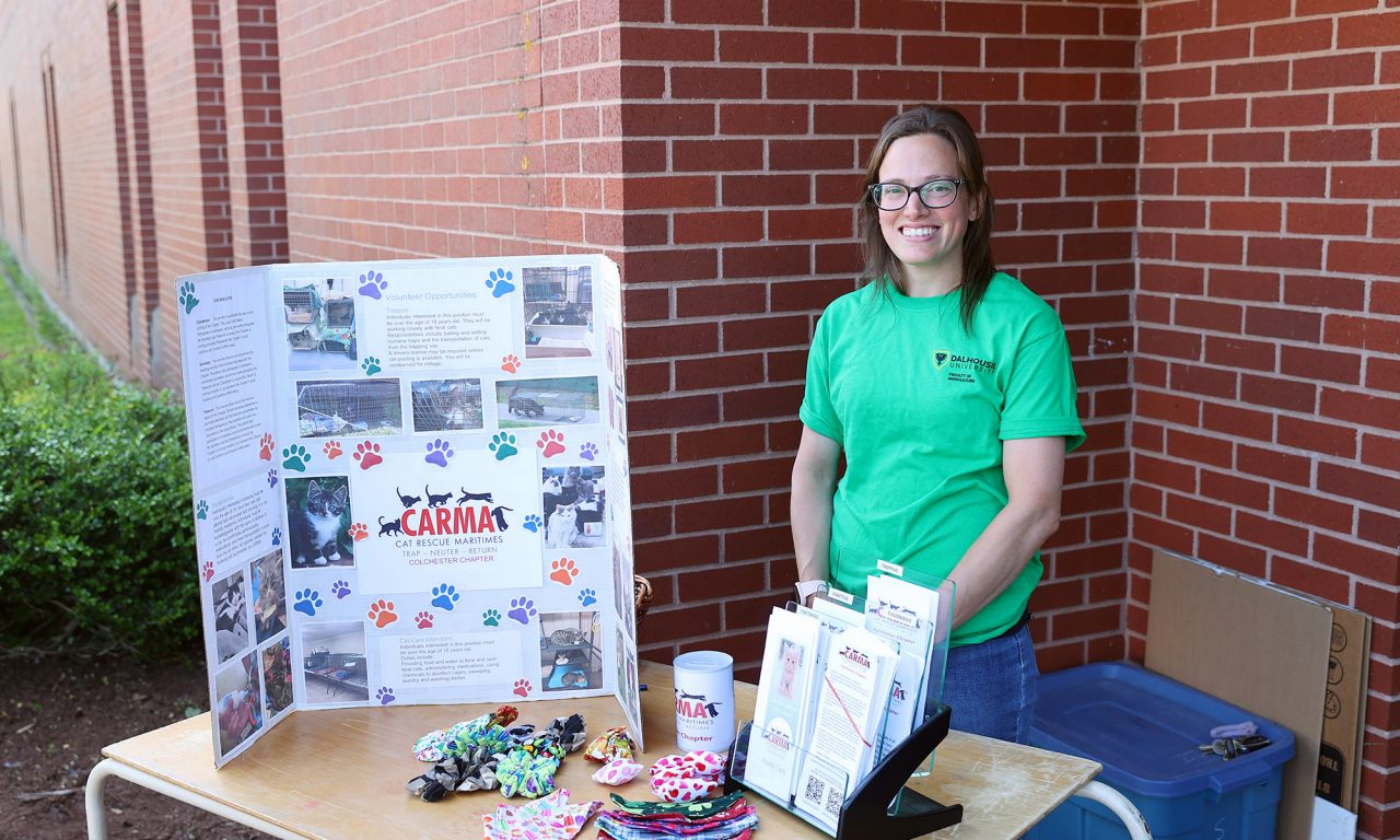 Woman with long dark hair and glasses wearing a bright green shirt stands behind a table displaying posters and brochures.