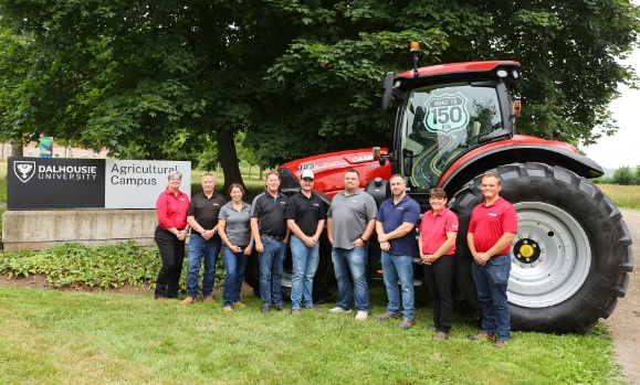 A group of nine individuals stands in a row in front of a large red tractor.