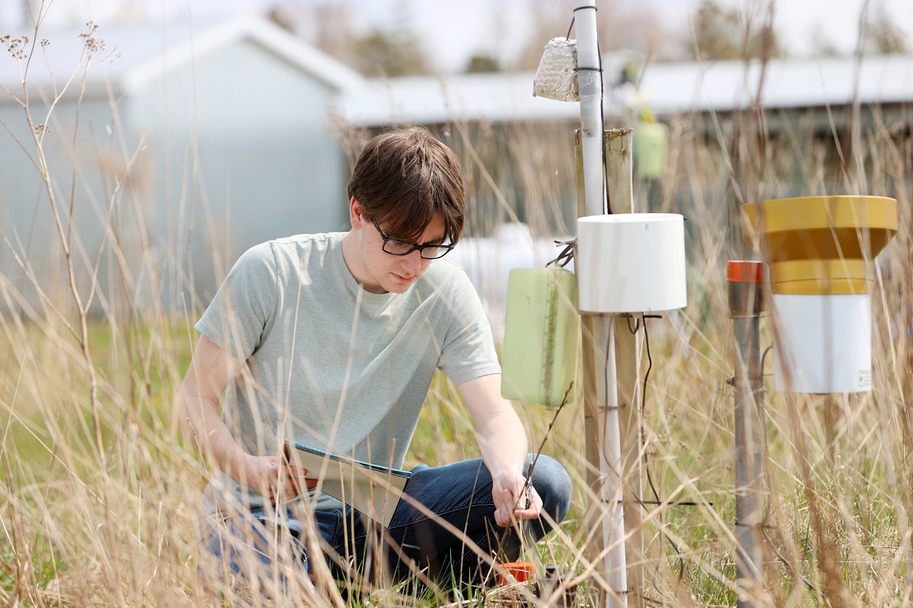 A student kneels in long grass in front of scientific equipment. They are wearing jeans and a t-shirt and holding a notepad.